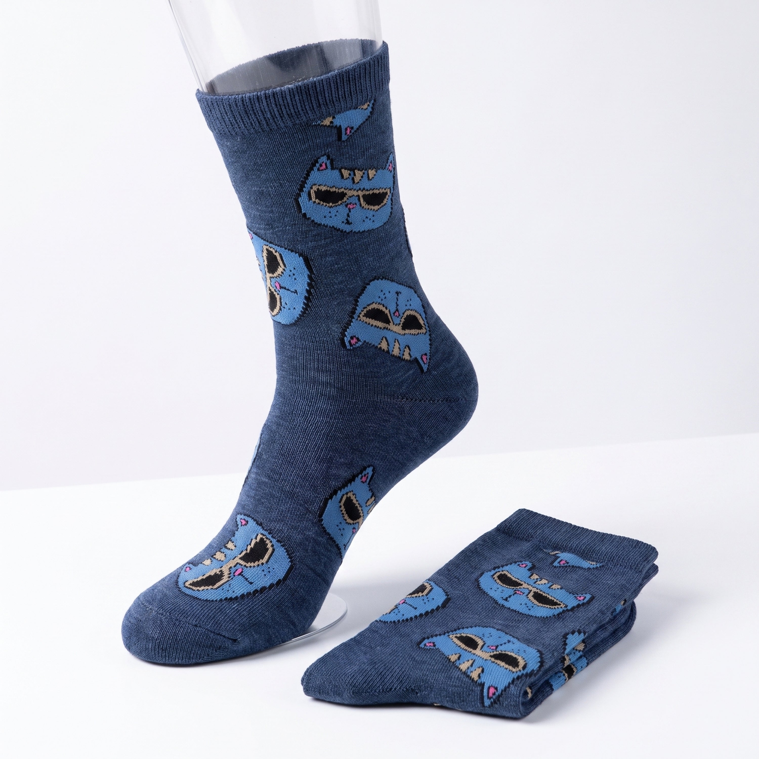 A pair of navy blue crew socks with a repeating pattern of blue cartoon monster faces, shown with one sock on a clear mannequin leg and the other folded on a white surface.