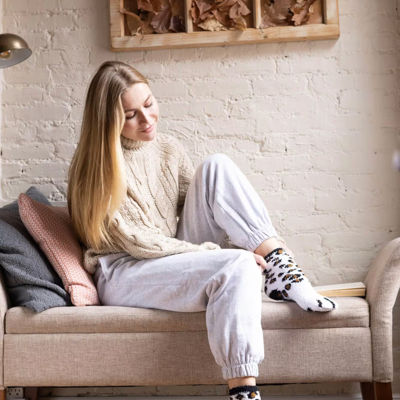 Woman sitting on a bench wearing a beige sweater, white pants, and patterned socks in a cozy room.