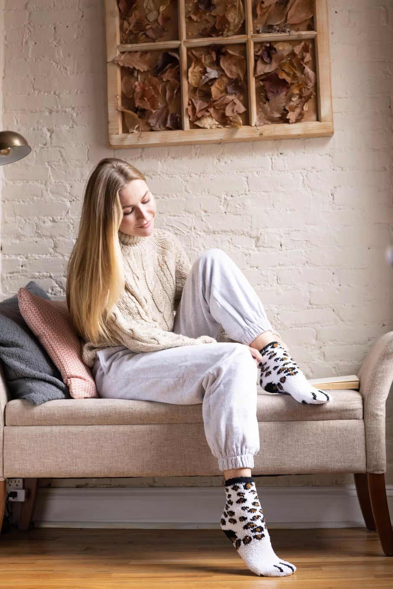Woman sitting on a bench wearing a beige sweater, white pants, and patterned socks in a cozy room.